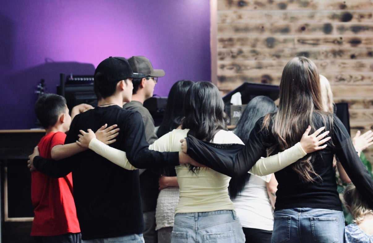 A group of people stands together with arms around each other, facing a purple-lit wall.