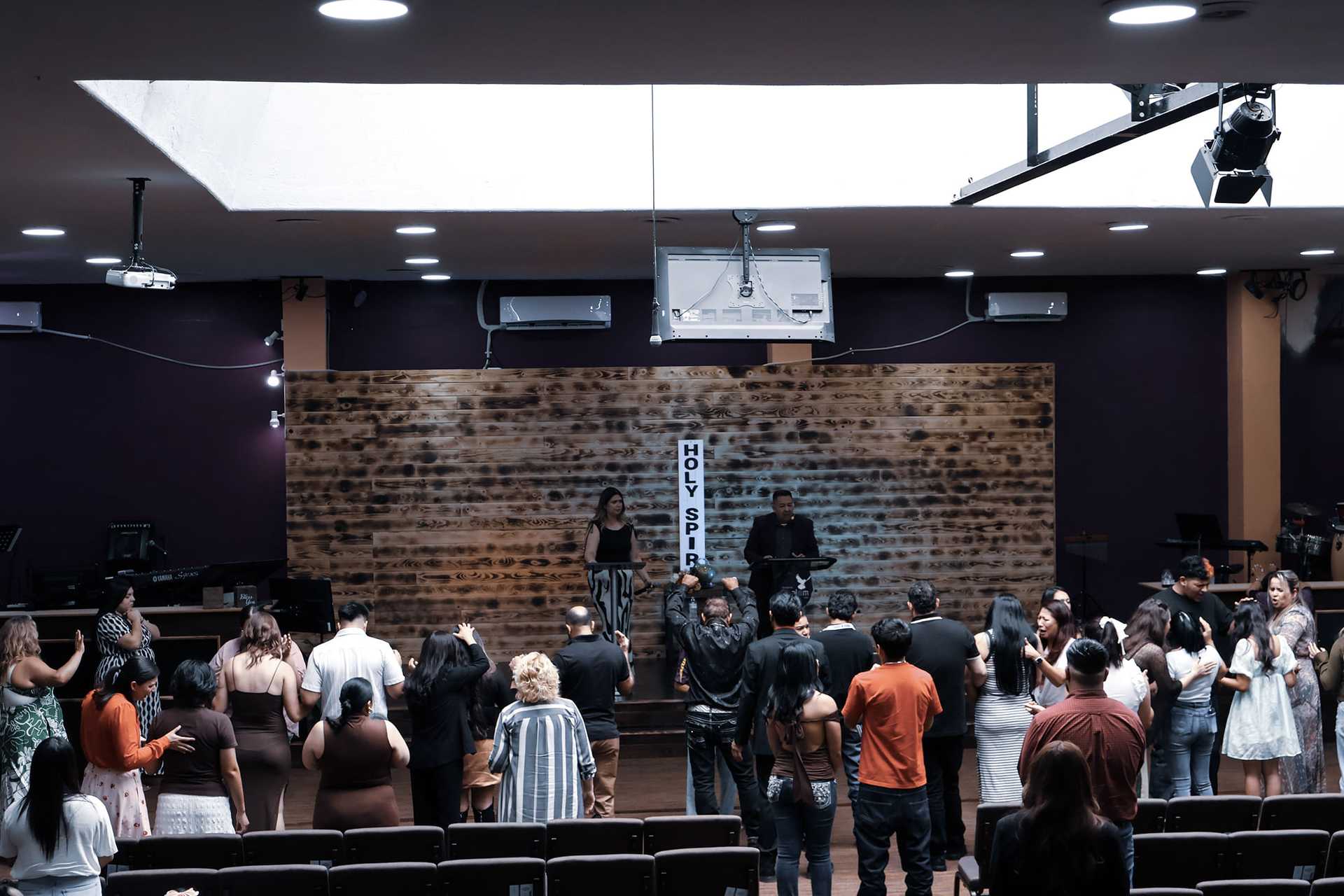 Congregation gathers in a church service, people standing and facing a stage with speakers.