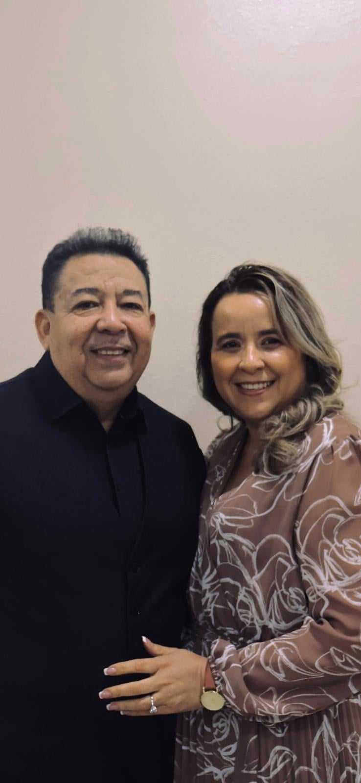 Man and woman smiling, standing close together against a light background. The woman is wearing a floral dress.