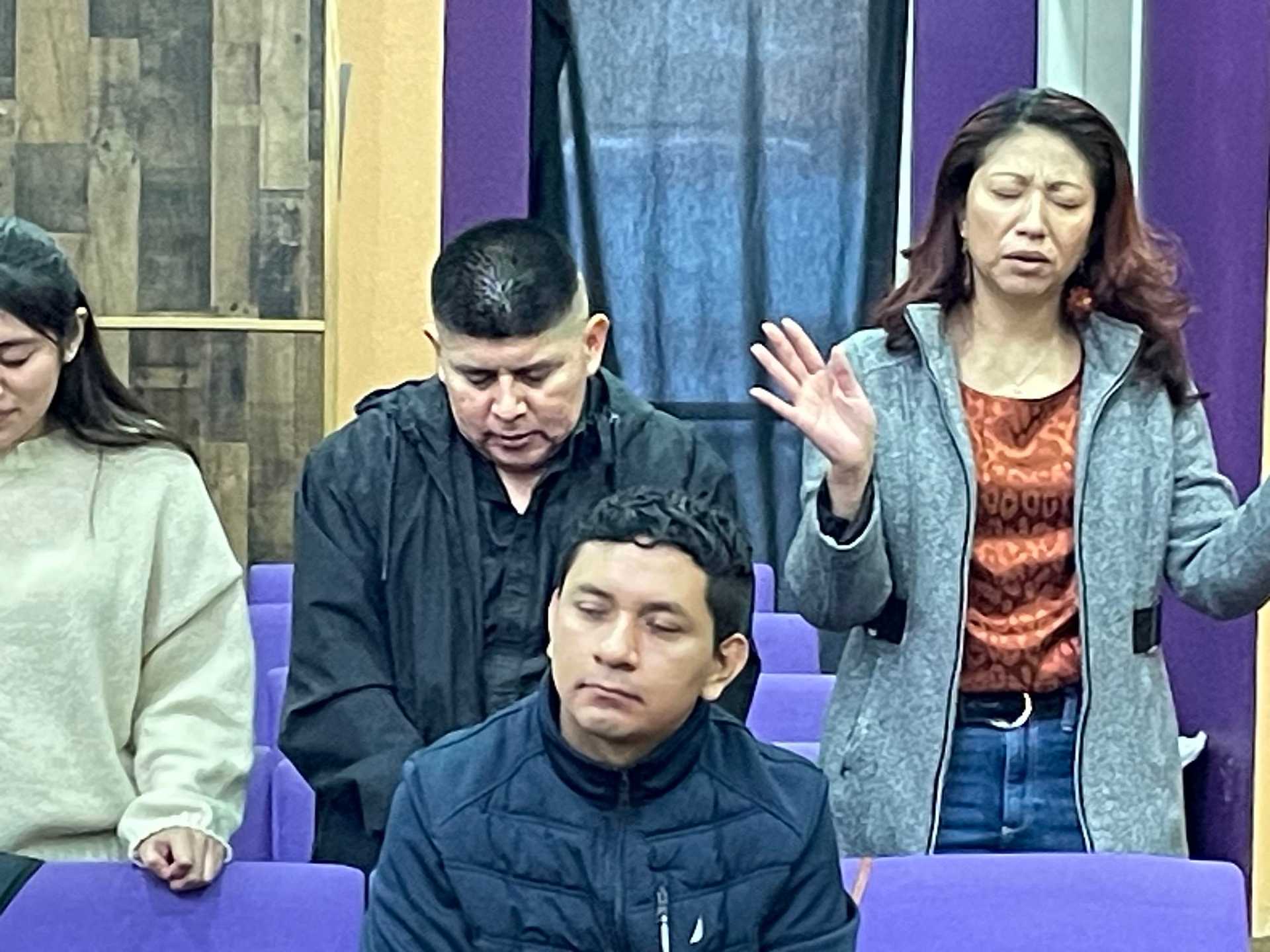 People in prayer at a church, standing and sitting with eyes closed and hands raised.