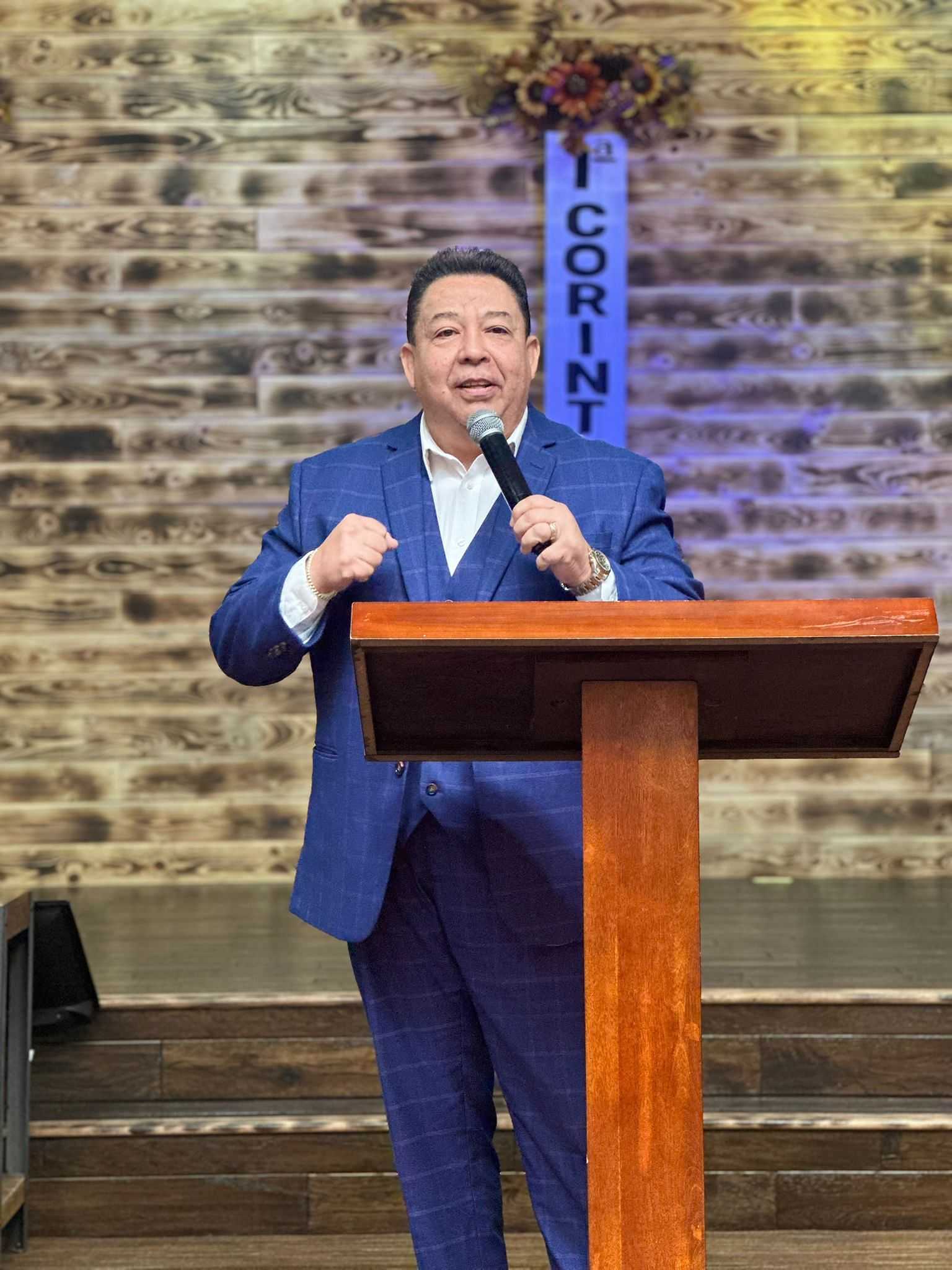 Man in a blue suit speaking at a wooden podium with "1 CORINTH" sign in the background.