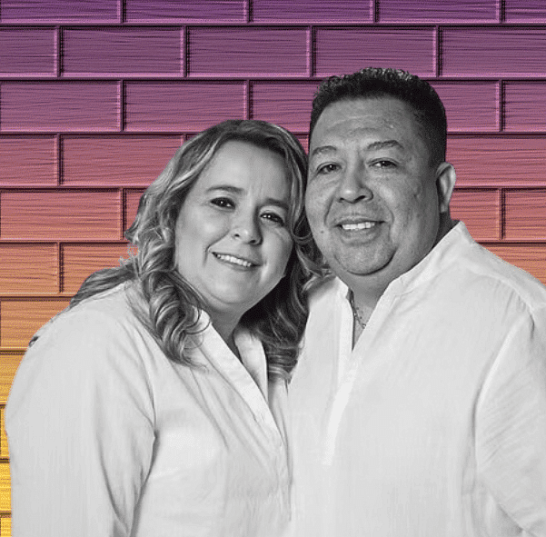 Smiling couple in white shirts stands together against a pink and orange brick wall background.