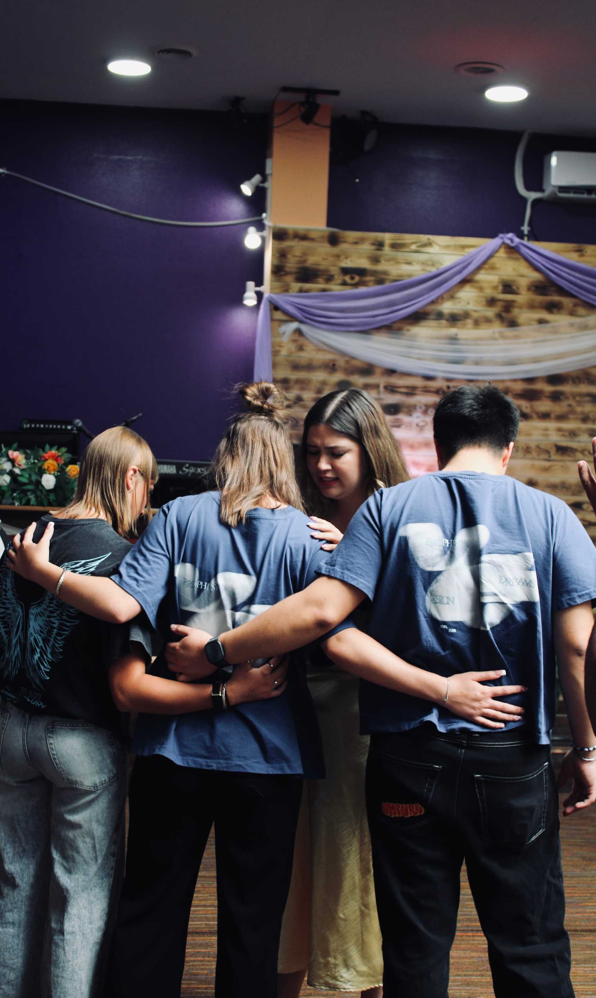 Group of friends embracing in a cozy room with purple walls and soft lighting.