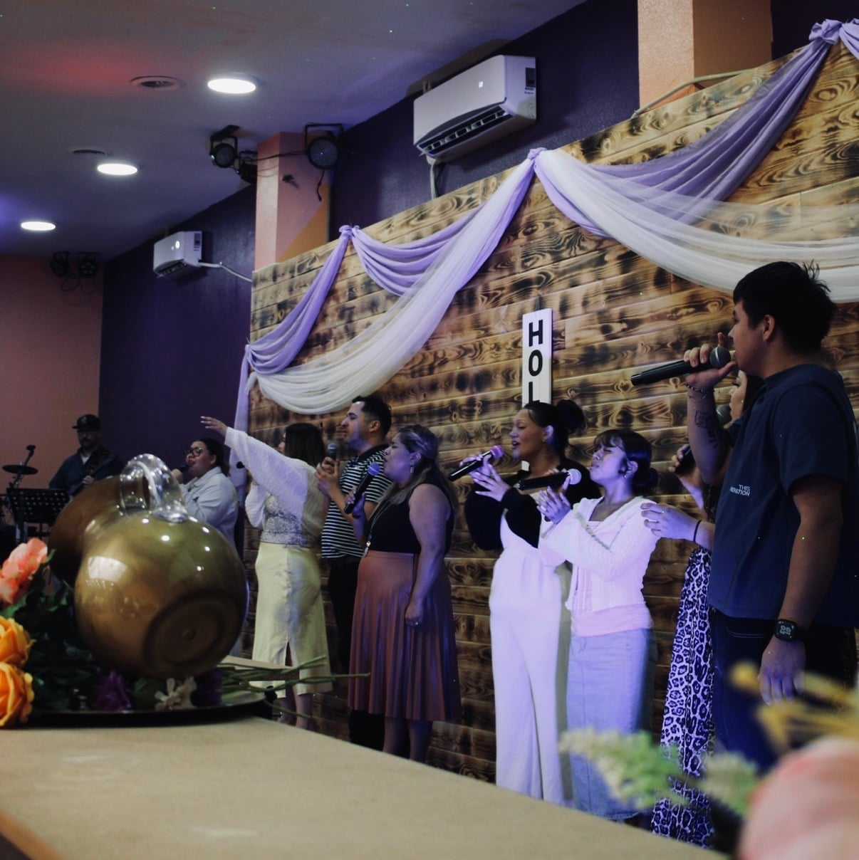 Group of people singing on stage together, decorated with drapes and floral arrangements.